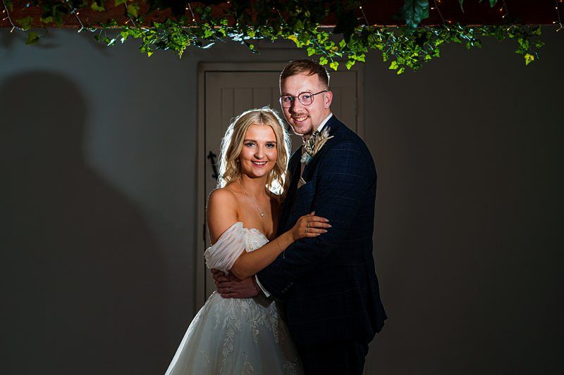 A couple in formal attire pose for a photo. The woman wears a white gown, while the man wears a dark suit with a boutonnière. They stand close together under a decoration of greenery. Image by Documentary Wedding Photographer Lewis Bishop Photography.