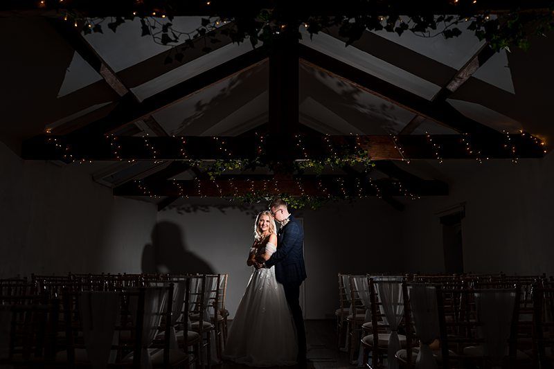 A bride and groom stand together in an empty, dimly lit hall adorned with string lights and wooden beams. Image by Documentary Wedding Photographer Lewis Bishop Photography.