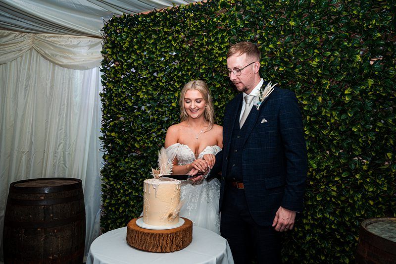 A bride and groom stand together, cutting a white frosted wedding cake placed on a wooden slab. They are in front of a leafy green backdrop, under a tent with draped fabric. Image by Documentary Wedding Photographer Lewis Bishop Photography.