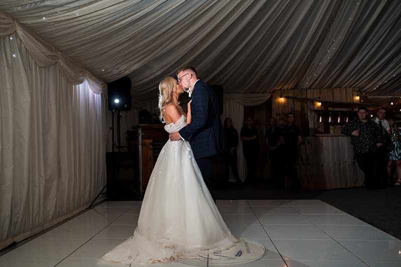 A bride and groom share their first dance in a tented venue; the bride is in a white gown and the groom is in a dark suit. The setting features draped white fabric and guests are visible in the background. Image by Documentary Wedding Photographer Lewis Bishop Photography.