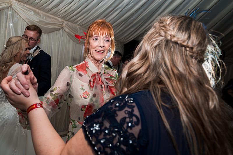 Two women are dancing together at an event, one in a floral dress and the other in a dark lace dress. A couple in the background is also dancing. Image by Documentary Wedding Photographer Lewis Bishop Photography.