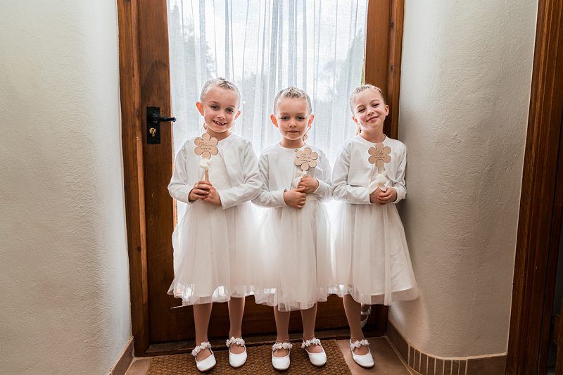 Three young children wearing white dresses and holding wooden clover-shaped signs stand in front of a door with a sheer curtain. Image by Documentary Wedding Photographer Lewis Bishop Photography.