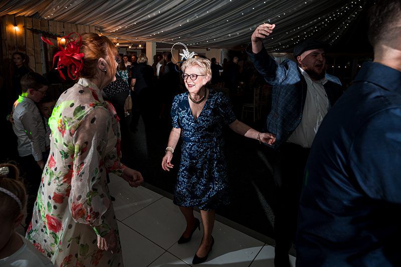 People dancing and socializing at an indoor event with festive lighting. Image by Documentary Wedding Photographer Lewis Bishop Photography.