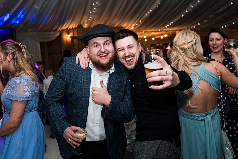 Two men at a lively event, both smiling and holding drinks, with one giving a thumbs-up. Guests in formal attire are visible in the background under string lights and a draped ceiling. Image by Documentary Wedding Photographer Lewis Bishop Photography.