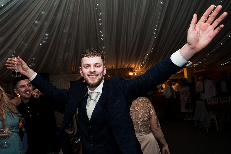 A man in a suit smiles and raises his arms in celebration at a lively event under a draped tent ceiling with string lights. Image by Documentary Wedding Photographer Lewis Bishop Photography.