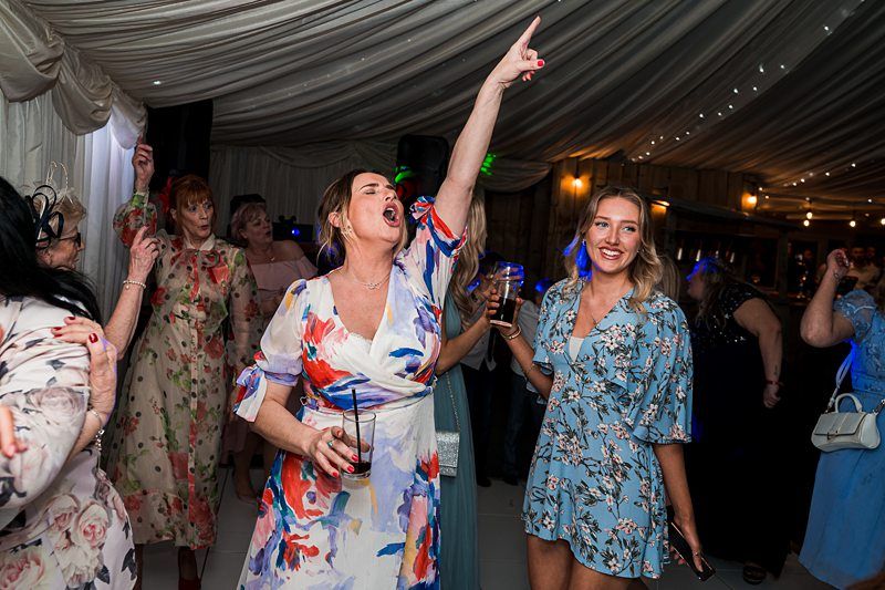 A group of women in colorful dresses dance and celebrate under a tent with string lights. One woman points upward and sings passionately while holding a drink. Image by Documentary Wedding Photographer Lewis Bishop Photography.