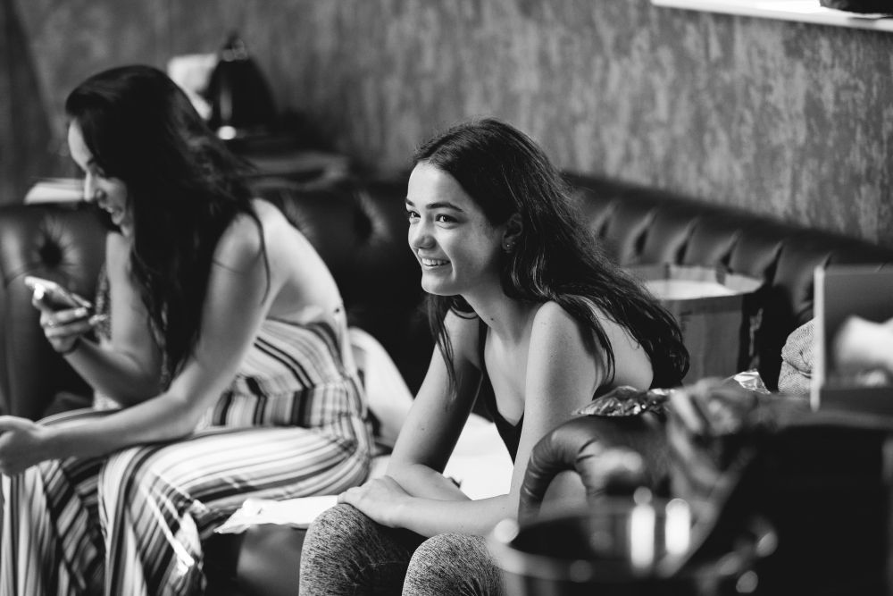 Black and white photo of two women indoors. One woman is sitting on a couch, smiling and looking ahead. The other woman is next to her, focused on her phone, and wearing a striped dress. The setting appears casual and relaxed, with some items visible on the side. Image by Documentary Wedding Photographer Lewis Bishop Photography.