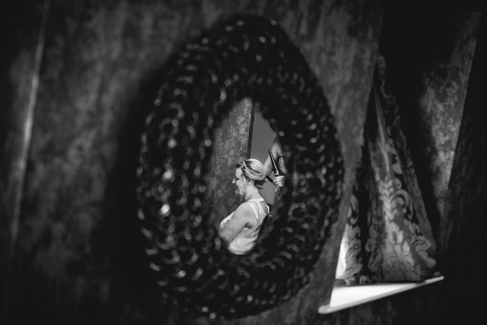 A black and white photo captures a woman reflected in an ornate oval mirror. She is seated with her arms crossed, looking away from the mirror. The background features patterned curtains and a textured wall. Image by Documentary Wedding Photographer Lewis Bishop Photography.