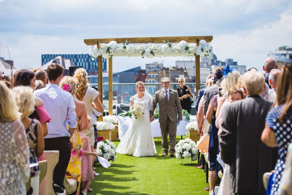 A bride in a white gown and a groom in a tan suit walk down an aisle outdoors under a wooden arch adorned with white flowers. Guests stand on either side, dressed in various styles, watching the couple proceed. It's a sunny day with a clear sky. Image by Documentary Wedding Photographer Lewis Bishop Photography.