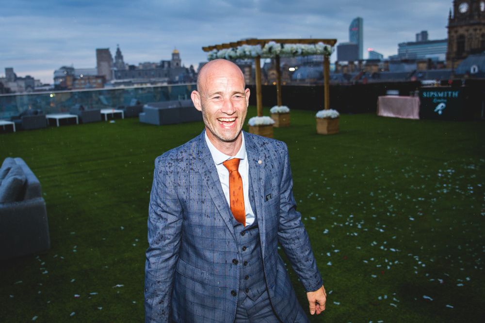 A bald man wearing a blue checkered suit with an orange tie is smiling and standing on a grassy area. It looks like it's raining as there are rain droplets on his suit. In the background, there is a wooden arch decorated with flowers, suggesting an outdoor event. Image by Documentary Wedding Photographer Lewis Bishop Photography.