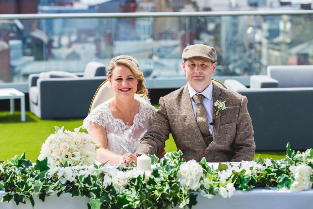 A newlywed couple sits at a table adorned with white flowers and greenery on a rooftop. The bride wears a lace dress and smiles, while the groom wears a tweed suit and a flat cap, looking ahead. The cityscape is visible in the background. Image by Documentary Wedding Photographer Lewis Bishop Photography.