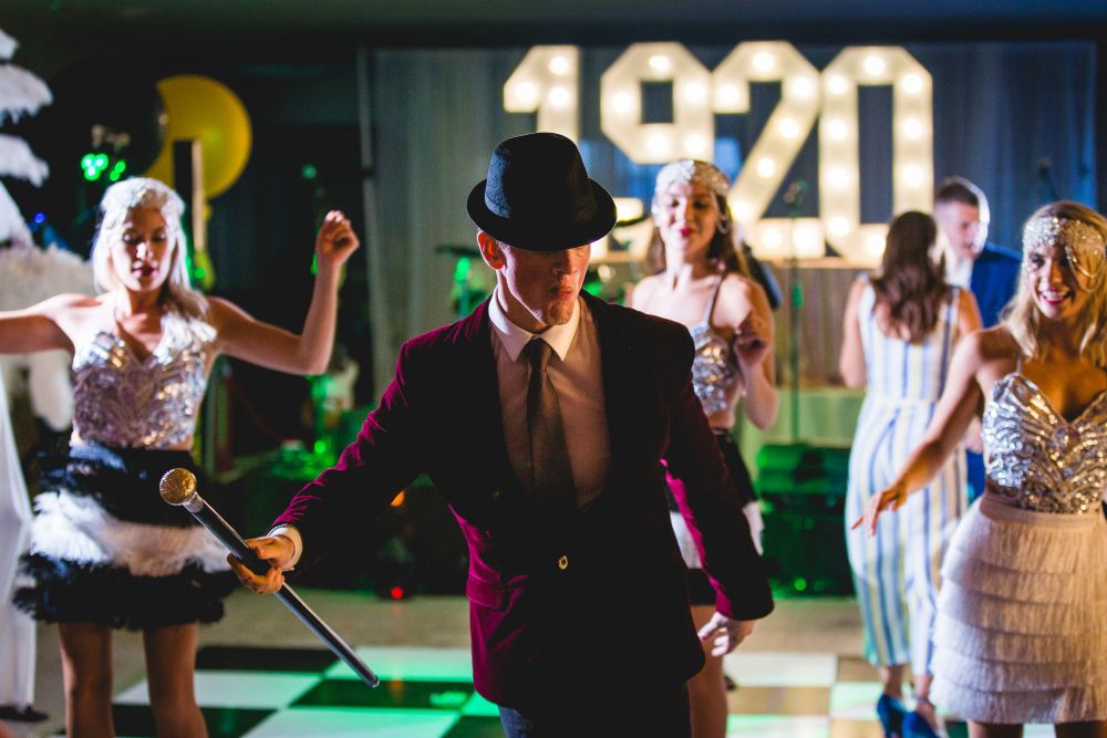 A person in a maroon jacket and black hat performs with a silver cane in front of a group of dancers dressed in sparkly costumes. In the background, large illuminated "1920" numbers and other dancers in vintage attire can be seen. Image by Documentary Wedding Photographer Lewis Bishop Photography.