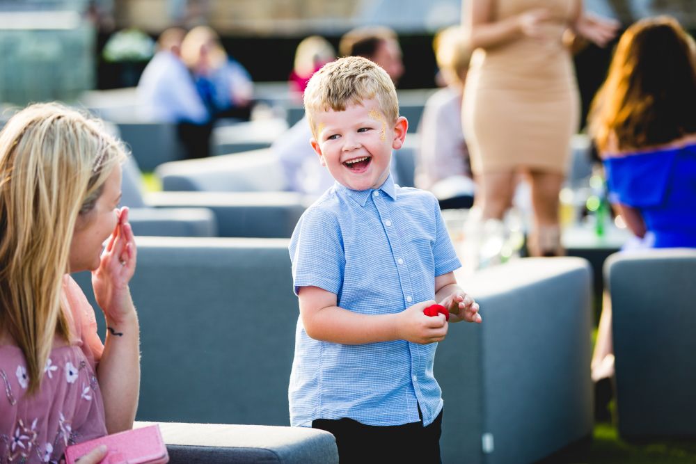 A young boy with blonde hair, wearing a light blue shirt, laughs joyfully while holding a small red ball in a relaxed outdoor social setting. People are seated on gray sofas in the background, engaged in conversation. Image by Documentary Wedding Photographer Lewis Bishop Photography.