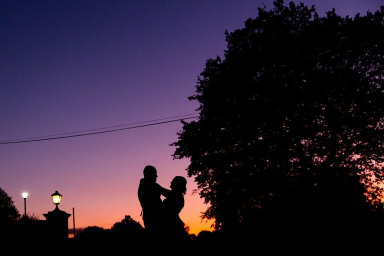 A silhouetted couple dancing under a tree at sunset, with a vibrant purple and orange sky. Streetlights are visible in the distance as the horizon darkens, casting a serene and romantic atmosphere. Image by Documentary Wedding Photographer Lewis Bishop Photography.