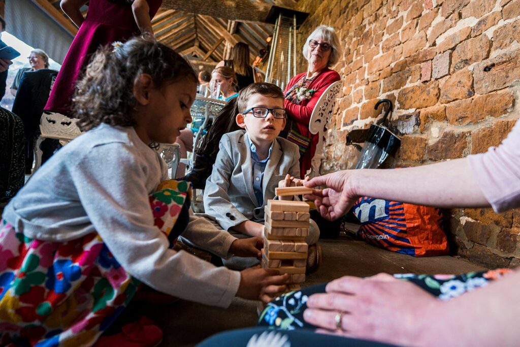 Children play with wooden blocks on the floor while adults look on in a room with exposed brick walls. Image by Documentary Wedding Photographer Lewis Bishop Photography.