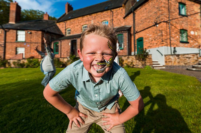 A boy with face paint smiles while leaning forward in front of brick houses on a sunny day. His hands rest on his knees, and another person is blurred in the background. Image by Documentary Wedding Photographer Lewis Bishop Photography.