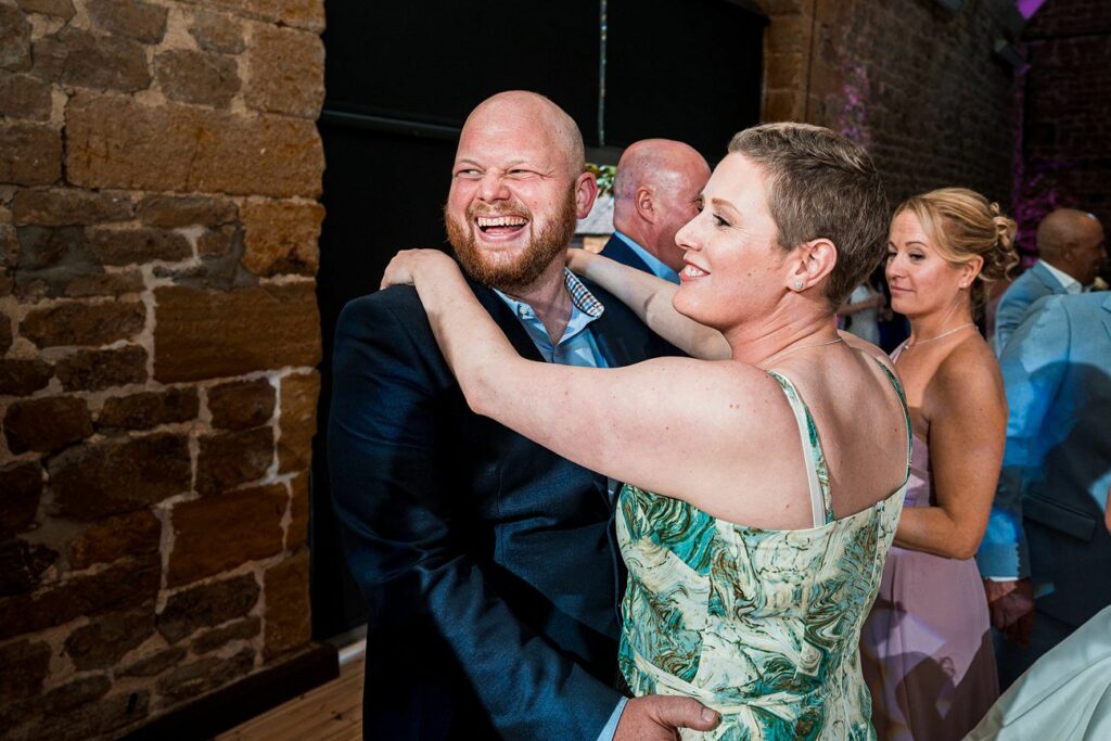 A man and woman share a cheerful moment while dancing together at an event, with other people dancing in the background. Image by Documentary Wedding Photographer Lewis Bishop Photography.