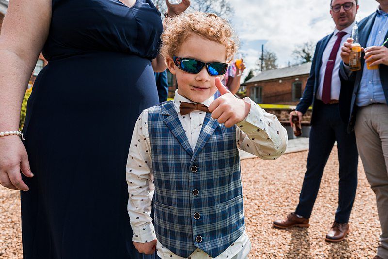 A young boy with curly hair wearing sunglasses and a plaid vest gives a thumbs up. Adults stand nearby holding drinks. Image by Documentary Wedding Photographer Lewis Bishop Photography.