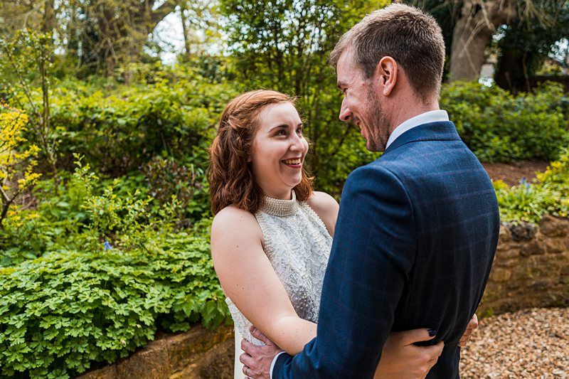 A woman and a man are standing close together outside, smiling and looking at each other. The background includes greenery and trees. Image by Documentary Wedding Photographer Lewis Bishop Photography.