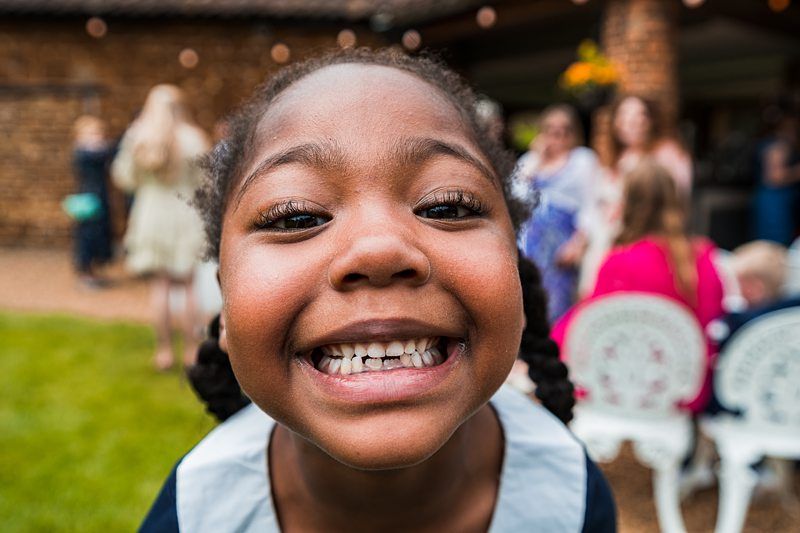 A close-up of a young child with braided hair smiling widely at the camera. There are people and outdoor furniture in the blurred background. Image by Documentary Wedding Photographer Lewis Bishop Photography.