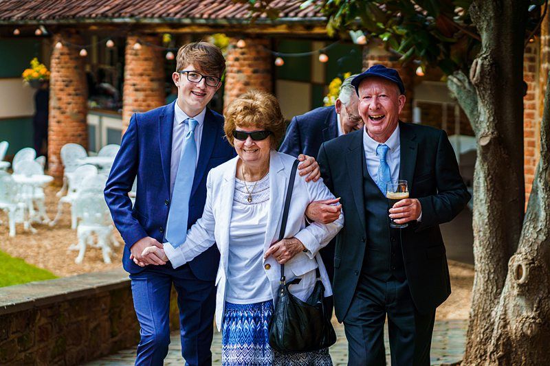An elderly man and woman, along with a young man, smile and walk arm-in-arm outdoors while dressed in formal attire. Image by Documentary Wedding Photographer Lewis Bishop Photography.