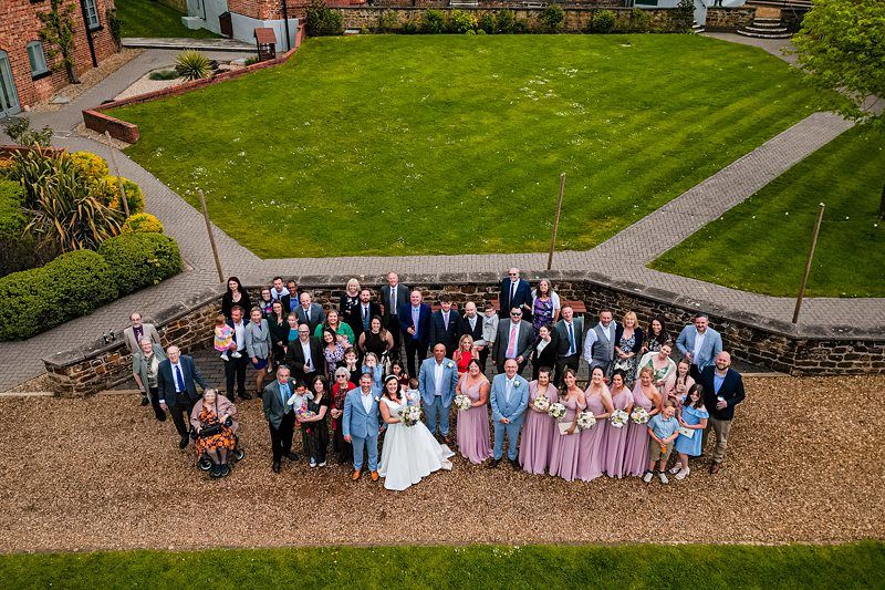 An aerial photo of a wedding party with about 50 people, including the bride, groom, bridesmaids, groomsmen, and guests, standing in a garden outside a rustic building with manicured lawns and pathways. Image by Documentary Wedding Photographer Lewis Bishop Photography.