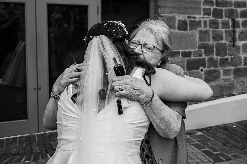 A bride in a wedding dress and veil embraces an elderly woman with glasses and a floral top outside a building with stone and brick walls. Image by Documentary Wedding Photographer Lewis Bishop Photography.