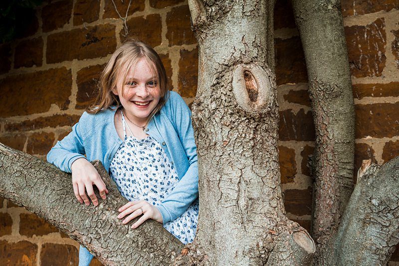 A young girl with blonde hair and a blue cardigan smiles while sitting in the branches of a large tree, with a stone wall in the background. Image by Documentary Wedding Photographer Lewis Bishop Photography.