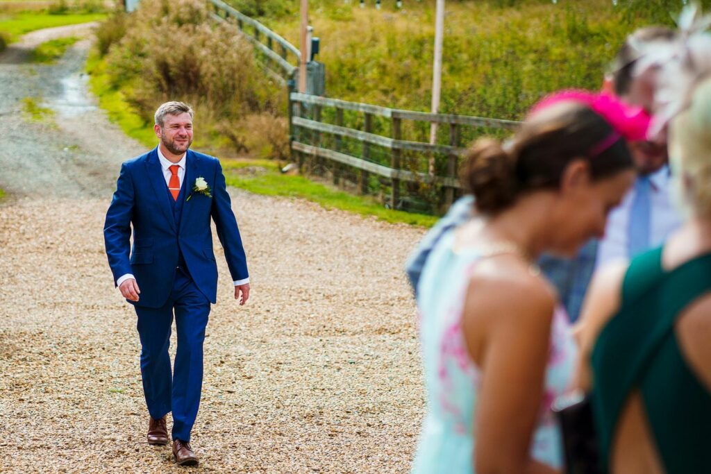A man dressed in a blue suit and orange tie, with a yellow boutonnière, walks on a gravel path towards a group of blurred people. The background features a rustic, wooden fence and green, grassy scenery. Image by Documentary Wedding Photographer Lewis Bishop Photography.