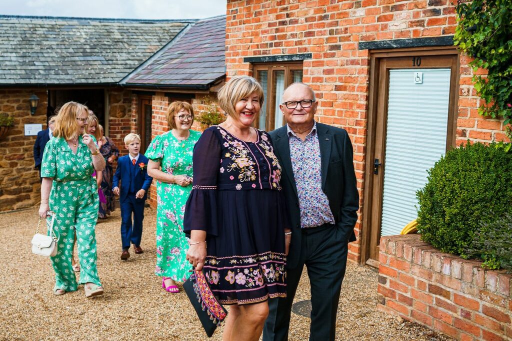 A group of people, smartly dressed, walk towards a brick building with numbered doors. The woman in the front wears a dark, floral dress and smiles at the camera, while the man beside her, in a suit, also smiles. Others in bright floral outfits follow behind. Image by Documentary Wedding Photographer Lewis Bishop Photography.
