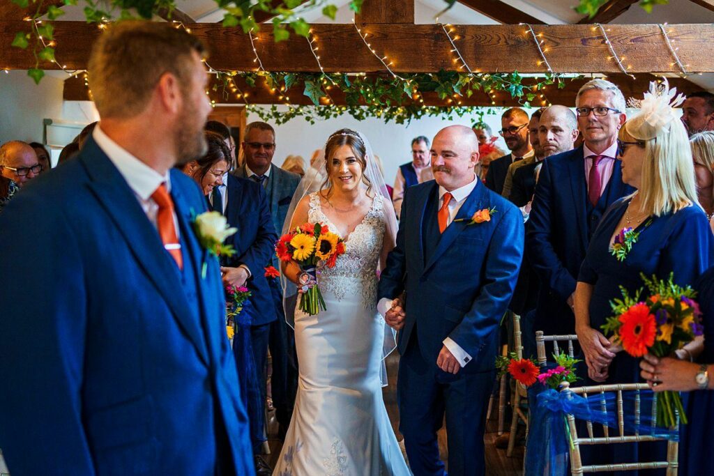 A bride holding a bouquet of orange and yellow flowers walks down the aisle with an older man. The aisle is decorated with greenery and string lights. The groom, dressed in a blue suit with an orange tie, waits at the end of the aisle, smiling. Guests look on. Image by Documentary Wedding Photographer Lewis Bishop Photography.