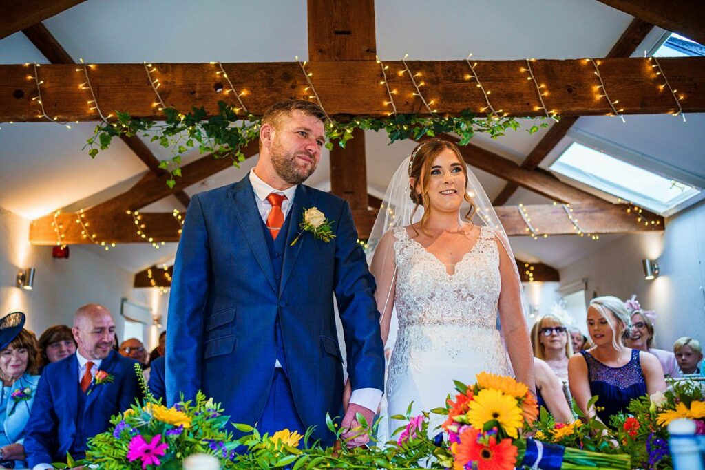 A bride and groom stand at the altar during their wedding ceremony. The groom wears a blue suit with an orange tie and boutonnière, and the bride wears a white lace gown and veil. They are surrounded by guests. Above them, fairy lights and greenery are draped along wooden beams. Brightly colored flowers are in the foreground. Image by Documentary Wedding Photographer Lewis Bishop Photography.
