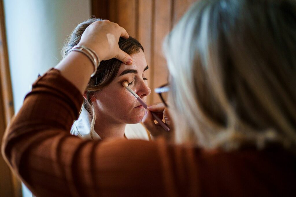 A woman with light brown hair is having her eyeshadow applied by another woman with blonde hair and glasses, who is using a makeup brush. The scene takes place indoors, possibly in preparation for an event such as a wedding. Image by Documentary Wedding Photographer Lewis Bishop Photography.