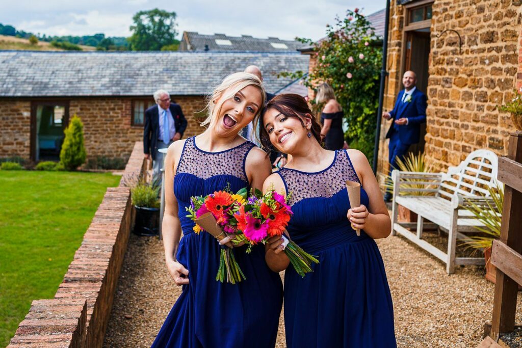 Two women in matching dark blue dresses pose playfully with colorful flower bouquets outside a rustic venue. One sticks her tongue out while the other smiles. They stand on a gravel path, with a stone building and lush greenery in the background. Image by Documentary Wedding Photographer Lewis Bishop Photography.