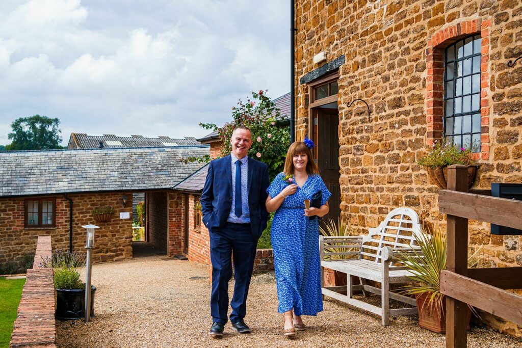 A man and woman, dressed in formal attire, walk along a gravel path beside a rustic brick building. The woman, in a blue dress, smiles and holds a drink. The sky is partly cloudy, and the scene exudes a cheerful, relaxed atmosphere. Image by Documentary Wedding Photographer Lewis Bishop Photography.
