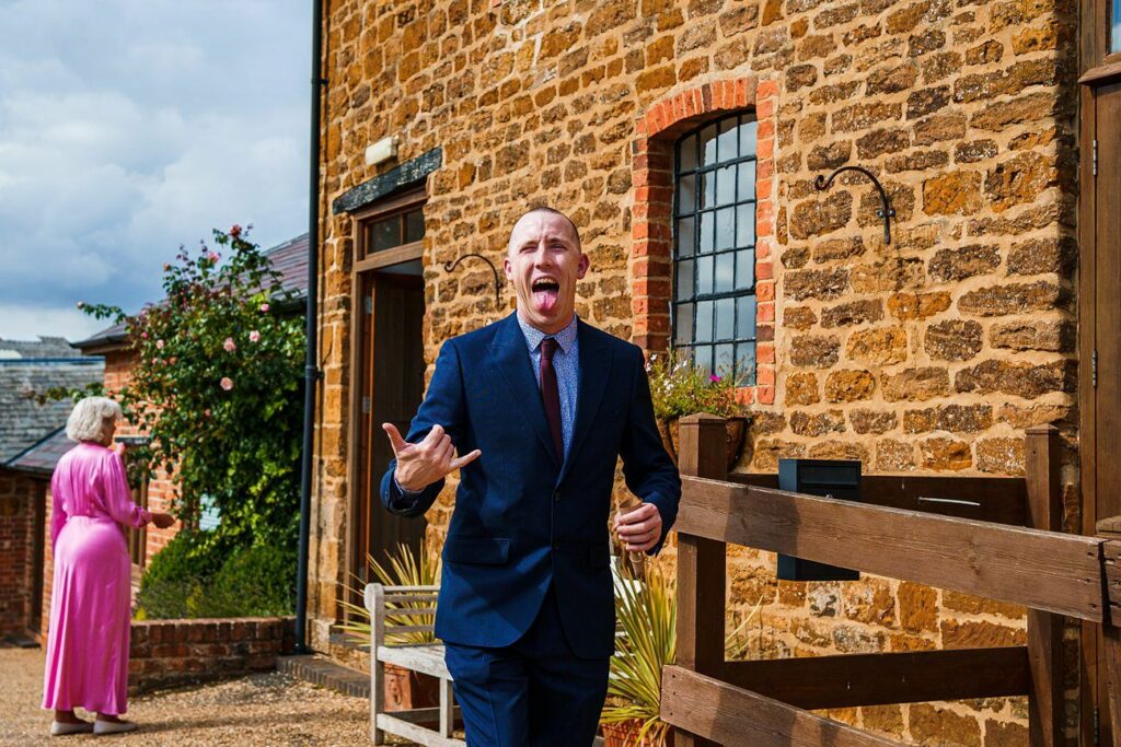 A man in a blue suit is playfully sticking out his tongue and pointing towards the camera while standing outside a rustic brick building. In the background, a woman wearing a pink dress walks along the pathway. The sky is partly cloudy. Image by Documentary Wedding Photographer Lewis Bishop Photography.