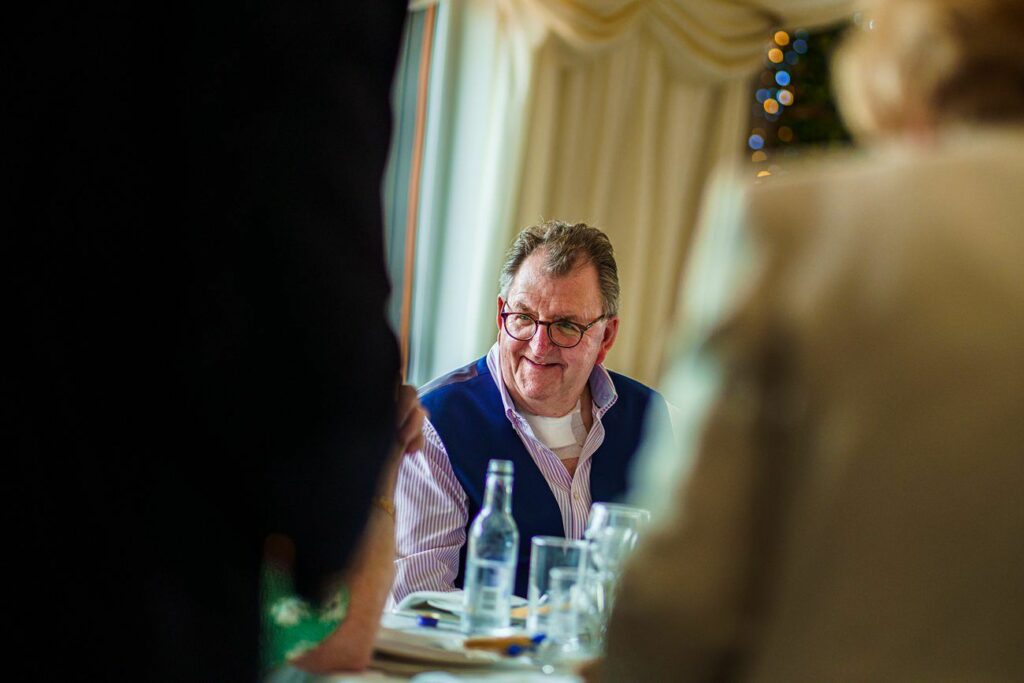 A man with glasses and a blue vest over a white shirt smiles while sitting at a table. Several water bottles and glasses are on the table, and other people are partially visible in the foreground. The background shows a window with curtains and some blurred decorations. Image by Documentary Wedding Photographer Lewis Bishop Photography.