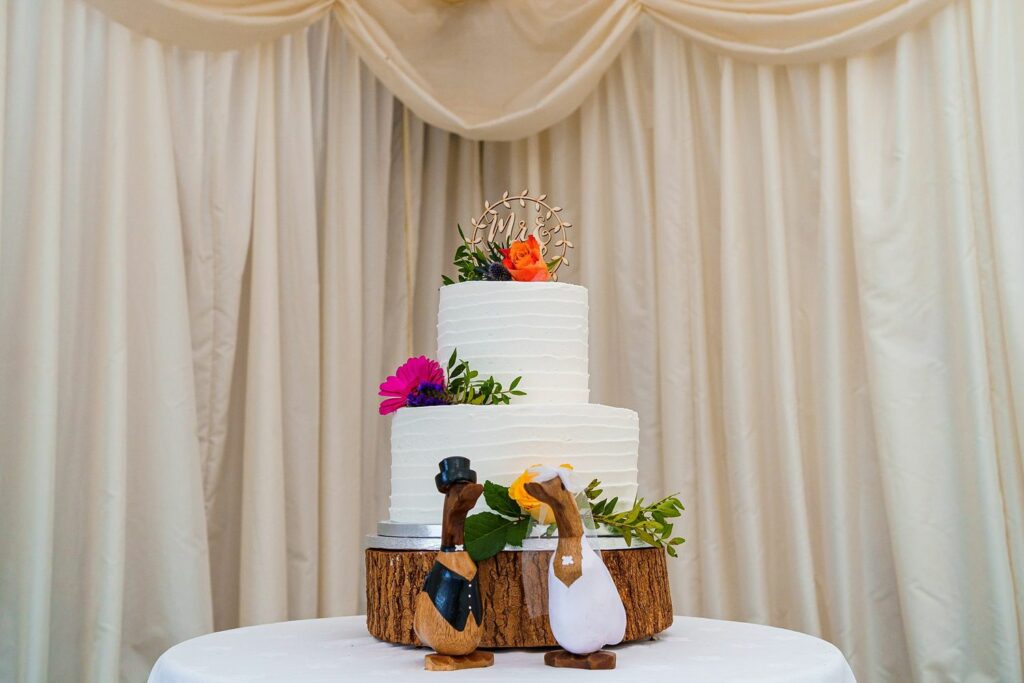 A three-tiered white wedding cake adorned with orange and pink flowers, green leaves, and a gold decoration sits on a wooden base on a round table. Two wooden duck figurines, dressed as a bride and groom, stand in front of the cake. Cream drapes form the backdrop. Image by Documentary Wedding Photographer Lewis Bishop Photography.