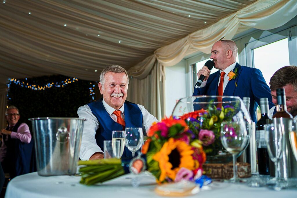 A man in a blue suit and orange tie sits at a table, smiling, while another man in a blue suit and holding a microphone speaks. The table has a colorful bouquet, wine glasses, a bucket, and various items. The room is decorated with lights and draped fabric. Image by Documentary Wedding Photographer Lewis Bishop Photography.