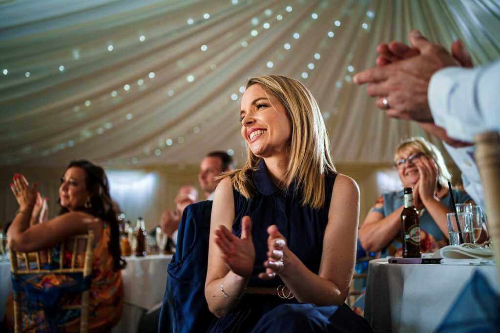 A woman in a dark dress sits at a table under a lit, draped canopy, smiling and clapping. Other guests around her, both men and women, are also clapping and appear to be enjoying themselves. The atmosphere is festive, suggesting a celebratory event. Image by Documentary Wedding Photographer Lewis Bishop Photography.