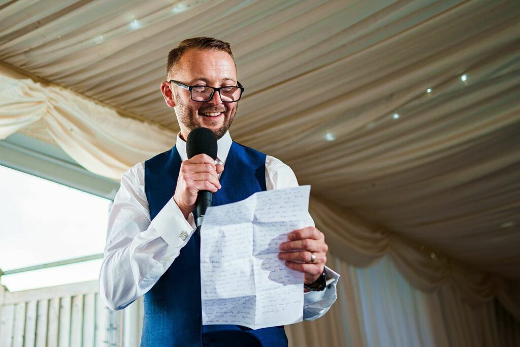 A man in a blue vest and white shirt is smiling while holding a microphone in his right hand and a handwritten speech in his left. He appears to be inside a decorated tent or hall, possibly delivering a speech at an event. Image by Documentary Wedding Photographer Lewis Bishop Photography.