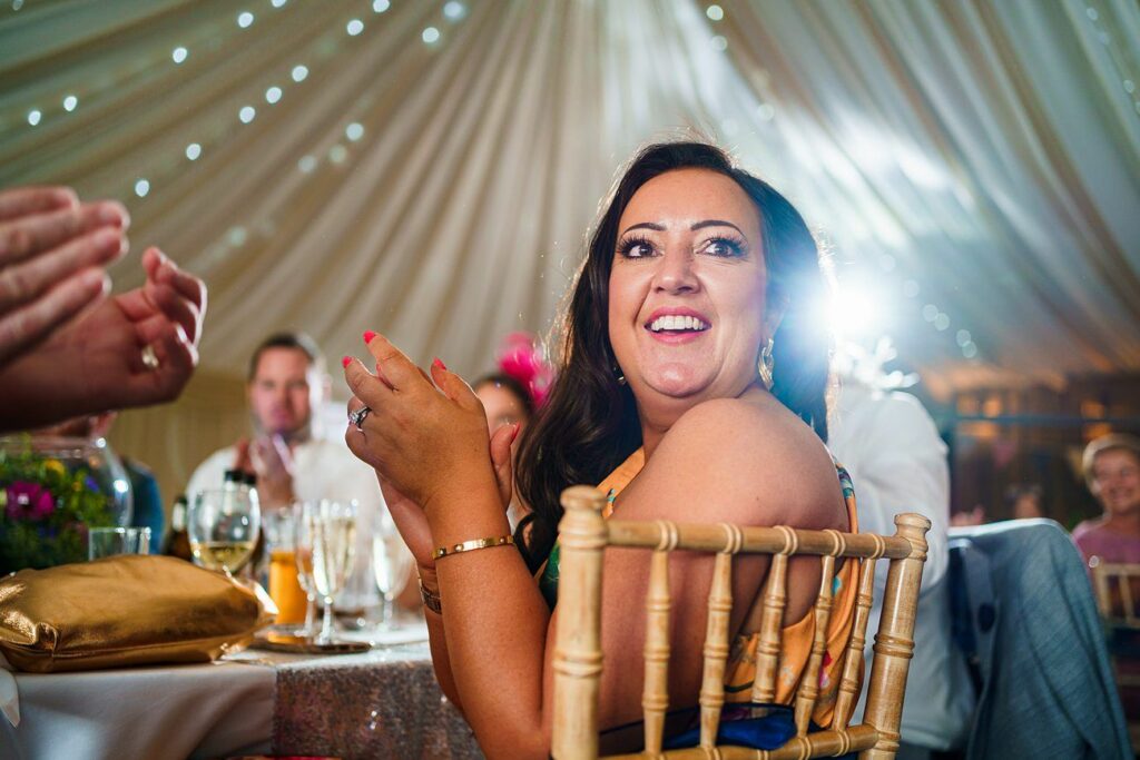 A woman with long dark hair, wearing a gold dress and jewelry, is smiling and clapping at an event. She sits at a decorated table inside a large, elegantly lit tent. Other people are visible in the blurred background, also clapping. Image by Documentary Wedding Photographer Lewis Bishop Photography.