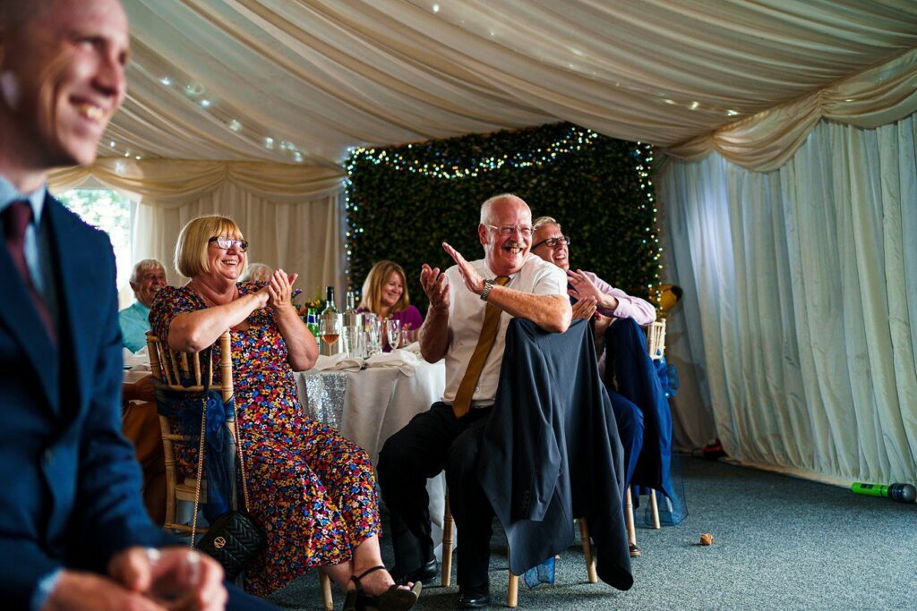 Guests in a decorated tent, including a man clapping and two women beside him smiling. The attendees are seated at round tables adorned with drinks and a sequined tablecloth. Fairy lights hang on the backdrop, adding a festive ambiance to the scene. Image by Documentary Wedding Photographer Lewis Bishop Photography.