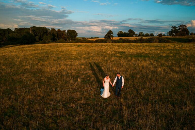 A bride in a white gown and a groom in a suit, holding hands, walk through a vast, open field at sunset. The sky is partly cloudy with a golden hue, and long shadows stretch behind them. Trees and a distant landscape frame the serene setting. Image by Documentary Wedding Photographer Lewis Bishop Photography.
