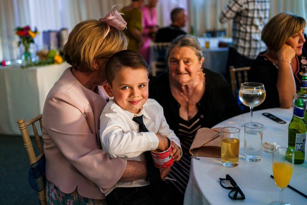 A young boy in a white shirt and black tie sits on a woman's lap at a table during a gathering. The boy is smiling and holding a small bucket. Two older women, one in a black top and another in a pink outfit with a fascinator, are seated nearby. Drinks are on the table. Image by Documentary Wedding Photographer Lewis Bishop Photography.
