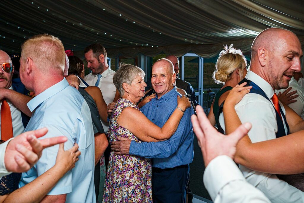 A group of people dance at a lively event under a tent with string lights. An older couple at the center smiles while dancing. Others dance around them, including a bald man in a vest and tie, and a man with red hair. The atmosphere is celebratory and joyful. Image by Documentary Wedding Photographer Lewis Bishop Photography.