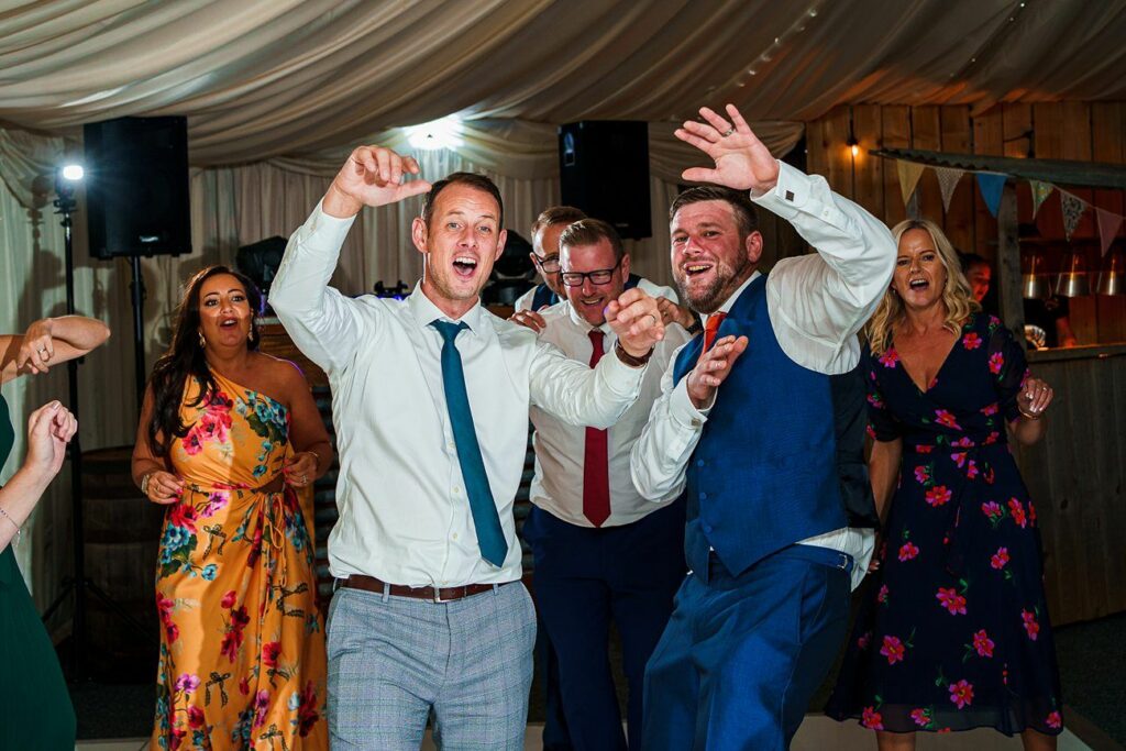 A group of people dressed in formal attire joyfully dance at a lively event under a tent with string lights. Two men in the front, one in a white shirt and blue tie, and the other in a blue suit, lead the group with animated expressions and raised arms. Image by Documentary Wedding Photographer Lewis Bishop Photography.