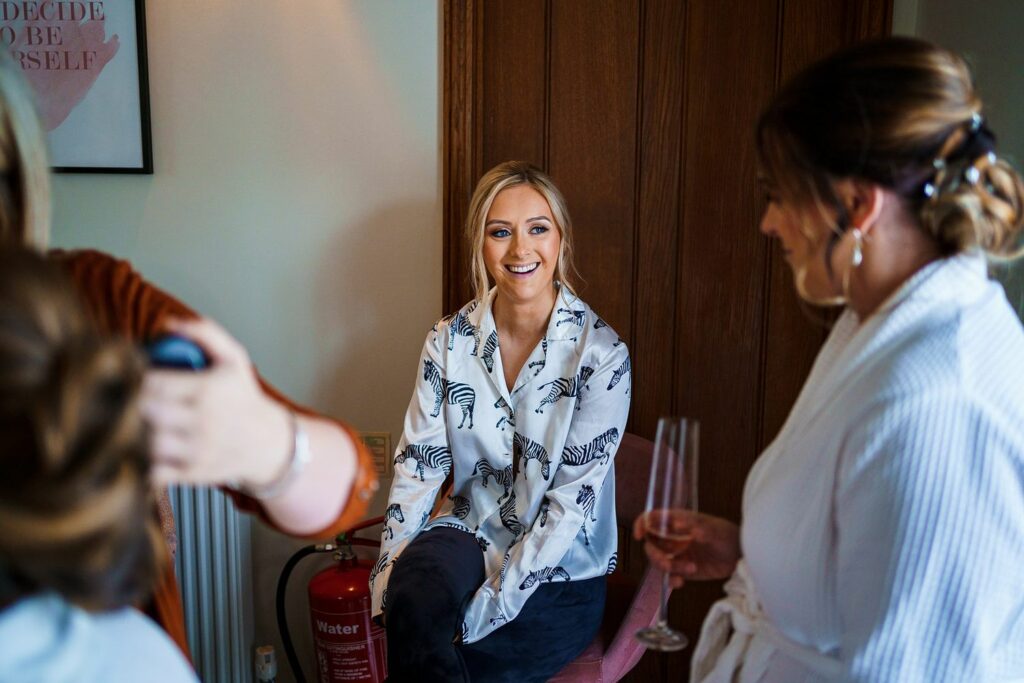 A woman in a patterned blouse sits on a chair, smiling, while talking to another woman in a robe holding a champagne glass. Another person off to the side appears to be styling hair. The setting is indoors with a wooden door and a framed picture on the wall in the background. Image by Documentary Wedding Photographer Lewis Bishop Photography.