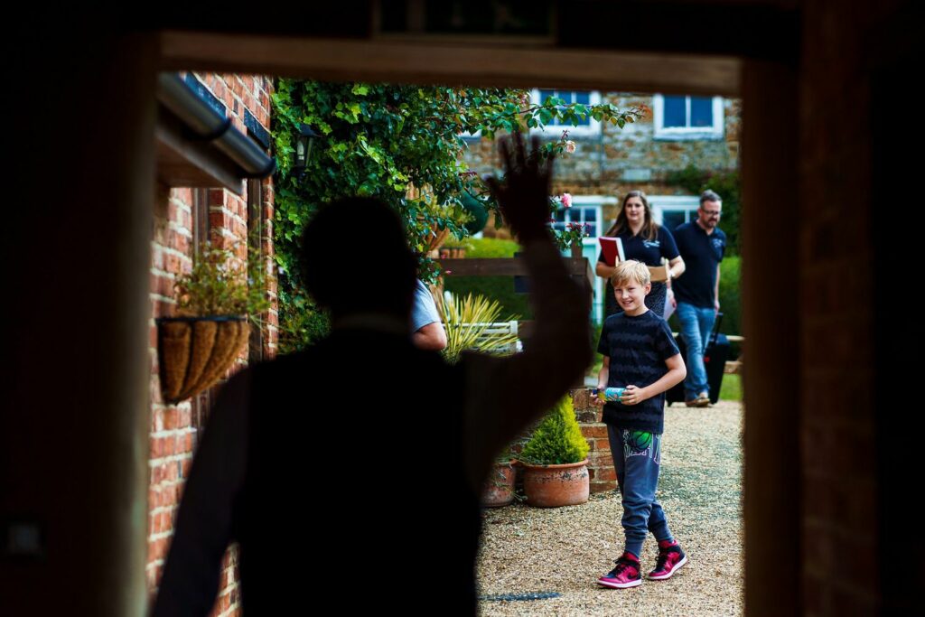 A person is waving while standing in a dark doorway. Outside, a child holding an object looks towards the door, and two adults in the background smile and walk. The setting seems to be a garden or outdoor area with plants, brick walls, and gravel path. Image by Documentary Wedding Photographer Lewis Bishop Photography.
