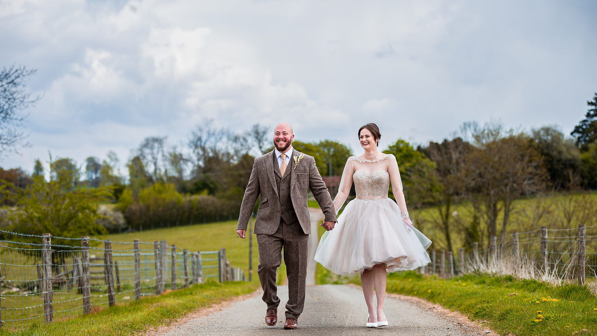 A smiling couple in wedding attire walks hand in hand down a country road. The groom wears a brown suit, and the bride wears a knee-length white dress. The background features lush green fields, trees, and an overcast sky. Image by Documentary Wedding Photographer Lewis Bishop Photography.